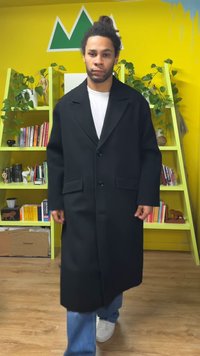 Abrigo largo negro con cierre de botones, grandes solapas y dos bolsillos frontales, hecho de una tela texturizada, usado sobre una camisa blanca y unos vaqueros.