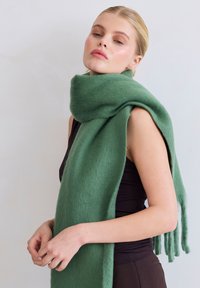 Groene wollen sjaal met een zachte textuur, voorzien van franjes aan de randen. Losjes om de hals gedragen, passend bij een donker shirt.