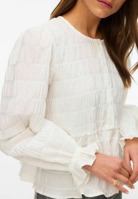 Femme portant un chemisier blanc texturé avec des manches longues bouffantes, une patte de boutonnage à l'avant et des poignets à volants, montrant ses mains et une partie de son visage.