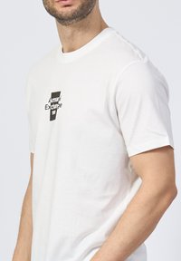Camiseta de algodón blanca con un logo gráfico negro. Corte estándar con mangas cortas y cuello redondo. Textura suave, diseño liso.