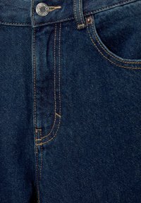 Gros plan d'un jean en denim bleu foncé montrant le bouton supérieur, la poche avant et les détails de couture beige.