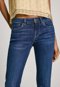 Jeans de denim azul oscuro con corte recto, que presentan costuras doradas, bolsillos delanteros y un cierre de botón. La tela tiene una textura suave.