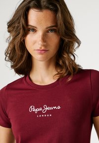 Bordová bavlnená tričko s okrúhlym výstrihom a krátkymi rukávmi, s bielym logom "Pepe Jeans London" potlačeným na hrudi. Hladká textúra.