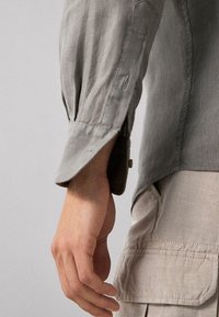 Linkerhand ontspannen langs de zijde hangend, gekleed in een lichtgrijze lange mouwen shirt en beige cargobroek met zichtbare klepzak.