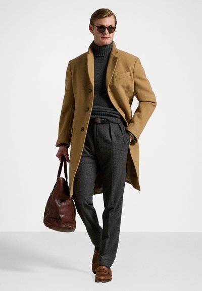 Brūns vilnas mētelis pār pelēku turtleneck, saskaņots ar pelēkiem biksēm un brūnām ādas kurpēm. Aksesuāri ietver brūnu ādas somu un saulesbrilles.