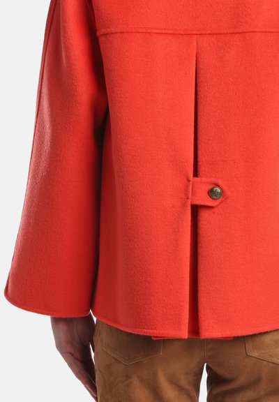 Raudonas vilnonis paltas su atvartais nugaroje, apvalia sagų užsegimu ir platėjančiomis rankovėmis. Dėvimas ant rudos spalvos zomšinėmis kelnėmis. Lygus tekstūros ir ryškios spalvos.