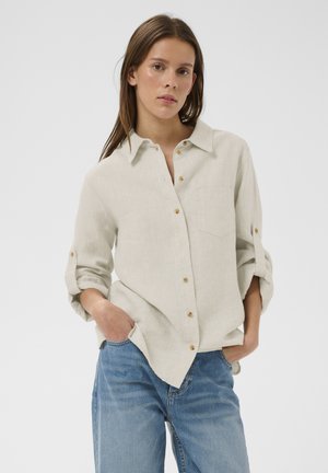 Młoda kobieta o długich brązowych włosach, ubrana w beżową koszulę na guziki z podwiniętymi rękawami oraz jasnoniebieskie dżinsy, ręce w kieszeniach, neutralne tło.