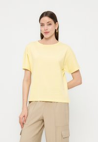 Νεαρή γυναίκα που φοράει ανοιχτό κίτρινο κοντομάνικο τοπ και μπεζ παντελόνι cargo, στέκεται με το ένα χέρι στην τσέπη μπροστά σε απλό λευκό φόντο.