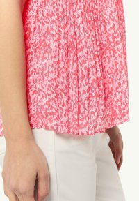 Różowa bluzka w wzory z plisowanym fasonem, wykonana z lekkiego materiału, zestawiona z białymi dopasowanymi spodniami. Zbliżenie na teksturę tkaniny.