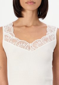 Valkoinen ribbattu tank top, jossa on monimutkainen pitsireunus kaula-aukossa ja harteilla, korostaen herkän kukkakuosin.