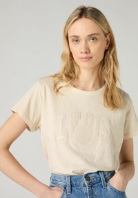 Vaalean beige puuvillapaita lyhyillä hihoilla, jossa on kohokuvioitu "LEVI'S" logo ja ympyrälogo edessä. Tyylitelty sinisillä farkkushorteilla.