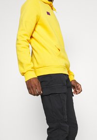 Geel hoodie met een voorzak, ribgebreide manchetten en een paarse merkembleem. Draagt met zwarte cargobroek met zijzakken.