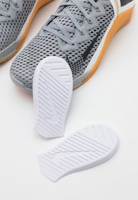 Scarpe sportive grigie con tomaia in rete, suola in gomma arancione e solette bianche con un motivo a righe in gomma e il logo Nike.