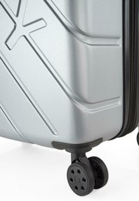 Maleta rígida plateada con líneas texturizadas, cremallera negra y dos ruedas negras con diseños perforados para estabilidad y maniobrabilidad.