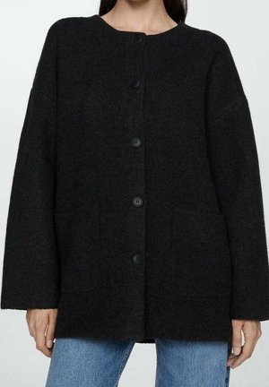Frau trägt eine schwarze, langärmlige Strickjacke mit Knöpfen und zwei Taschen vorne, kombiniert mit hellblauen Jeans.