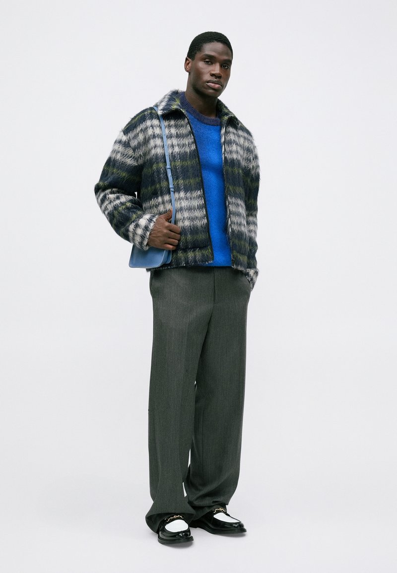 Hombre con chaqueta de pelaje estampado, suéter azul brillante, pantalones anchos grises, mocasines en blanco y negro, y bolso azul cruzado, de pie contra un fondo blanco.