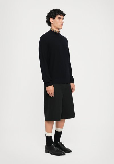 Maglione a coste navy con colletto a righe colorate, abbinato a pantaloni corti neri fino al ginocchio e scarpe stringate nere con calzini bianchi.