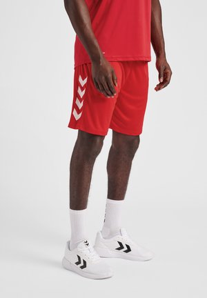 Rode sportieve shorts met een witte chevronontwerp aan de zijkant, gecombineerd met witte schoenen met zwarte accenten en witte sokken.