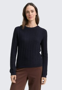 Marineblauer gerippter Pullover mit langen Ärmeln und Rundhalsausschnitt, aus weichem Material gefertigt. Kombiniert mit braunen Hosen für einen Kontrast.