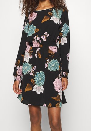 Robe noire à manches longues avec imprimé floral bleu, rose et blanc, taille élastique et poignets smockés, portée par une femme aux cheveux bouclés.