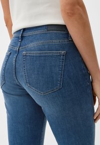 Blaue Denim-Jeans haben eine mittlere Taille, ein Fünf-Taschen-Design, gestickte Akzente und eine glatte Textur. Das Markenschild ist am Bund sichtbar.