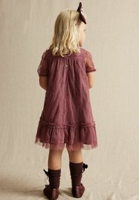 Bordeauxrotes besticktes Kleid mit kurzen transparenten Ärmeln, gerafftem Saum und Knopfverschluss. Kombiniert mit braunen knielangen Socken und Samtschuhen.