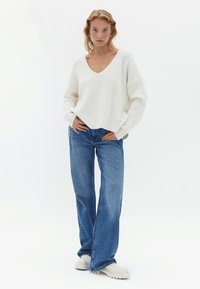 Witte gebreide trui met een V-hals, gecombineerd met blauwe wijde jeans en witte chunky schoenen. De trui heeft een geribbelde textuur.