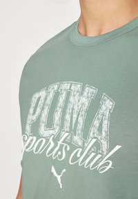 Grünes Baumwoll-T-Shirt mit einem großen weißen Grafik, auf der "PUMA Sports Club" steht. Glatte Textur mit klassischem Rundhalsausschnitt.