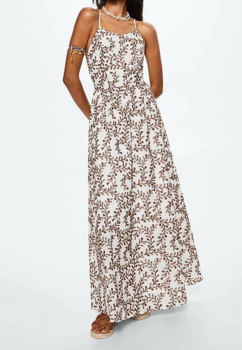 Femme portant une robe maxi blanche sans manches avec un motif de feuilles marron, associée à des accessoires de cou et de bras perlés et des sandales marron.