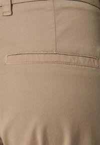 Dettaglio della tasca in tessuto beige con cuciture dritte, caratterizzato da un design a fessura orizzontale. Materiale texture con finitura liscia.