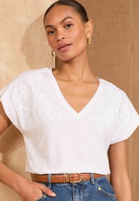 White V-neck t-shirt with short sleeves, featuring floral embroidery on the shoulders and relaxed fit. Paired with high-waisted denim jeans.