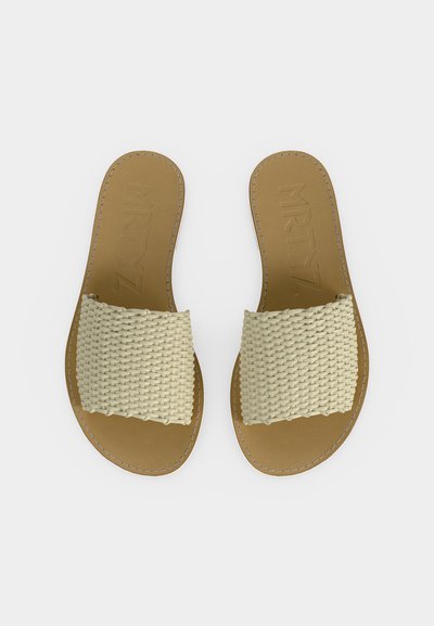 Ζευγάρι επίπεδων μπεζ σανδαλιών με φαρδιές πλεκτές κρεμί λουριά, ορατή ραφή κατά μήκος των σόλων και ανάγλυφο κείμενο στις εσωτερικές σόλες.
