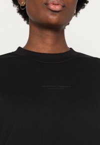 Černé bavlněné tričko s vroubkovaným kulatým výstřihem a diskrétním potiskem na přední straně. Tkanina je hladká a mírně přiléhavá.