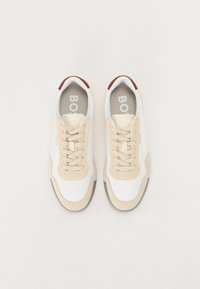 Hvite og beige sneakers med teksturert overflate, med en kontrasterende rød hæl, flate snører og en polstret innersåle merket med "BOSS."