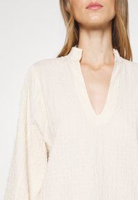 Femme portant un chemisier texturé de couleur crème avec un col en V et un petit col montant, visible des épaules au menton sur fond blanc.