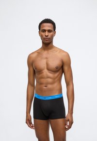 Boxer briefs neri con una fascia blu che presenta il logo "TOMMY HILFIGER". Il modello ha una corporatura snella ed è in piedi contro uno sfondo neutro.