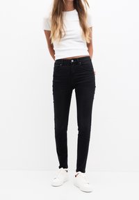 Jean skinny noirs en denim, avec poches avant, fermeture à bouton et une silhouette ajustée, associés à un T-shirt blanc et des baskets.