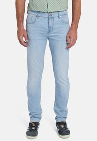 Jeans in denim azzurro chiaro, slim fit, cinque tasche, leggera scoloritura, dettagli cuciti, chiusura con bottone e cerniera, indossati con sneakers blu navy.
