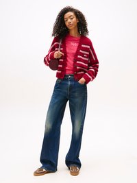 Cardigan en tricot rayé rouge et rose, chemise rose avec logo, jeans bleu ample et baskets marron avec accents blancs. Sac à main rond.