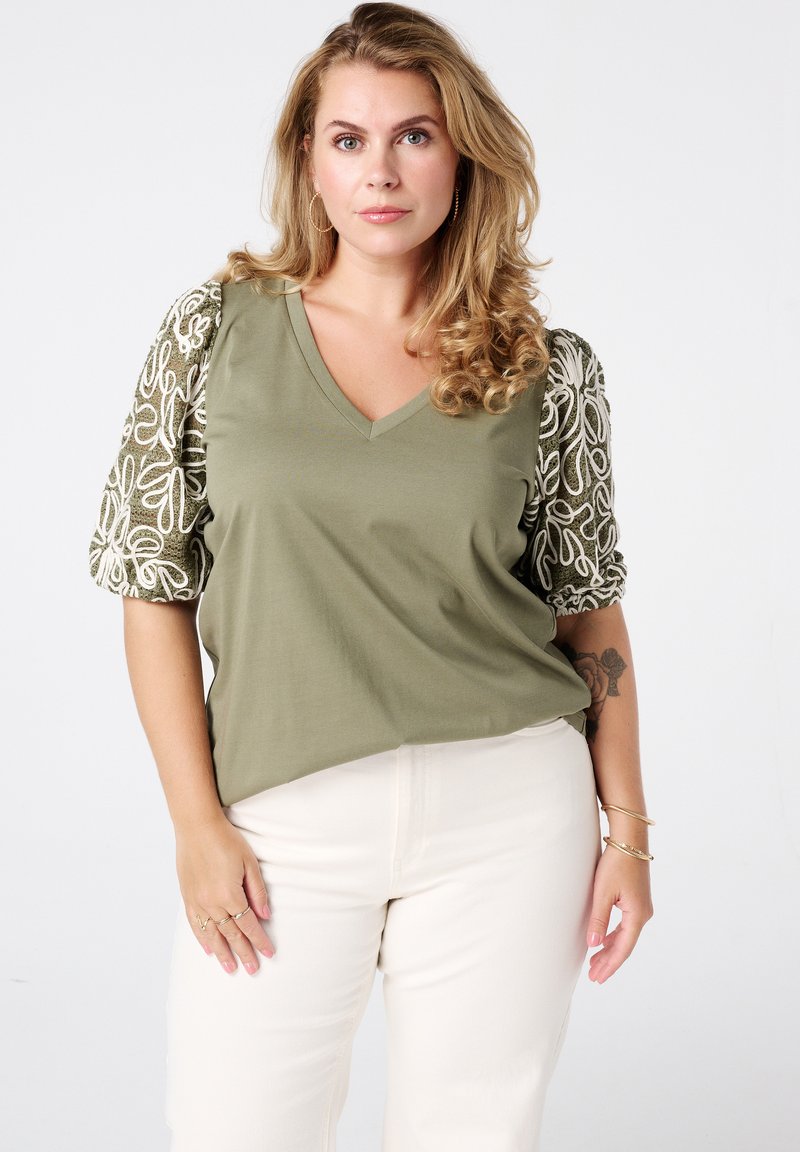 Olivengroene top met een V-hals en korte, gestructureerde mouwen met witte bloem borduursels. Draagt met lichtgekleurde broek.