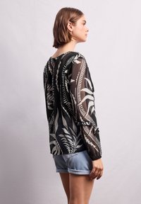 Giovane donna in blouse trasparente nera con motivi di foglie bianche e pantaloni corte di denim chiaro arrotolati, rivolta verso destra.