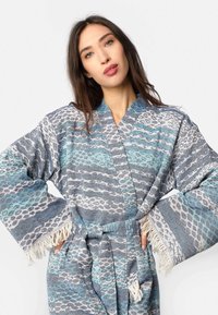 Ženska z dolgimi rjavimi lasmi nosi modro in sivo vzorčasto haljo z resastimi rokavi, roke ima na bokih, pred enobarvno belo kuliso.