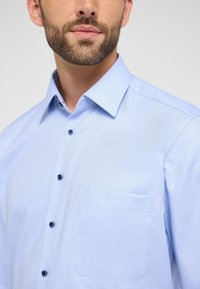 Lichtblauw overhemd van stof met textuur, een button-down kraag, zijzak en donkerblauwe knopen.