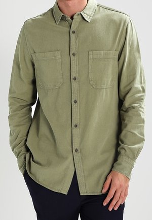 Mand iført en olivengrøn skjorte med lange ærmer og knapper, to brystlommer og sorte bukser, stående med den ene hånd i lommen.