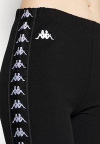 Czarne legginsy z szerokim elastycznym pasem i pionowymi białymi akcentami logo wzdłuż zewnętrznych szwów. Miękka, elastyczna tkanina.
