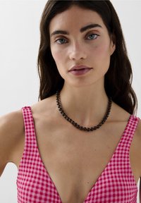 Zwarte kralenketting met ronde kralen, gedragen bij een roze ruitjesjurk met een diepe V-hals en brede schouderbanden.