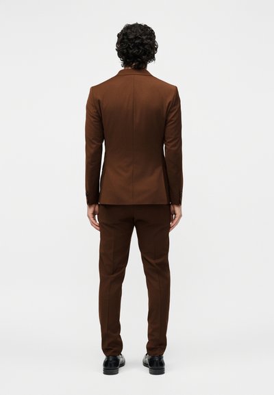 Άνδρας με σγουρά σκούρα μαλλιά, γυρισμένος με την πλάτη, φοράει μια εφαρμοστή καφέ στολή και μαύρα επίσημα παπούτσια σε ένα απλό φωτεινό φόντο.