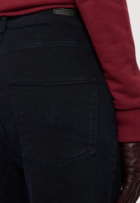 Tmavě modré džíny s texturovaným povrchem, na zadním kapsu s embosovaným logem. V kombinaci s vínovým dlouhým rukávem a hnědými rukavicemi.