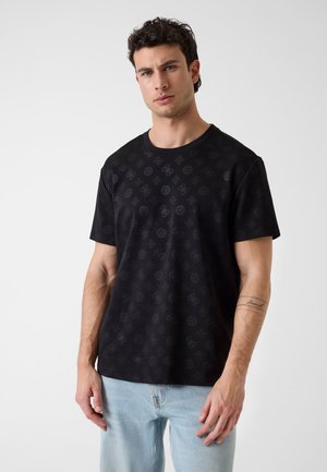 Jonge man met kort krullend haar, gekleed in een zwart T-shirt met patroon en lichtblauwe jeans, staand tegen een effen witte achtergrond.