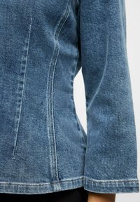 Jasnoniebieska kurtka dżinsowa z widocznymi detalami szwów. Charakteryzuje się dopasowanym krojem i długimi rękawami, teksturowany materiał z lekkim wytarciem.